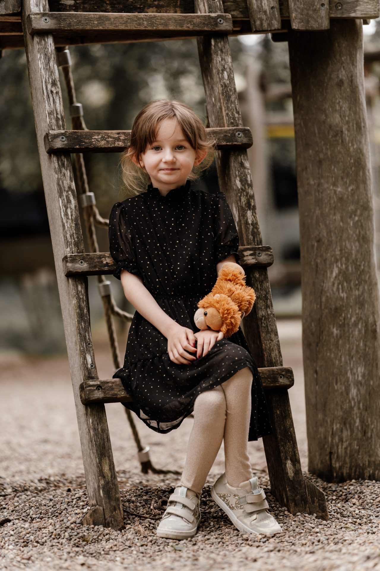 Mädchen sitzt mit Kuscheltier auf einer Holzleiter am Spielplatz