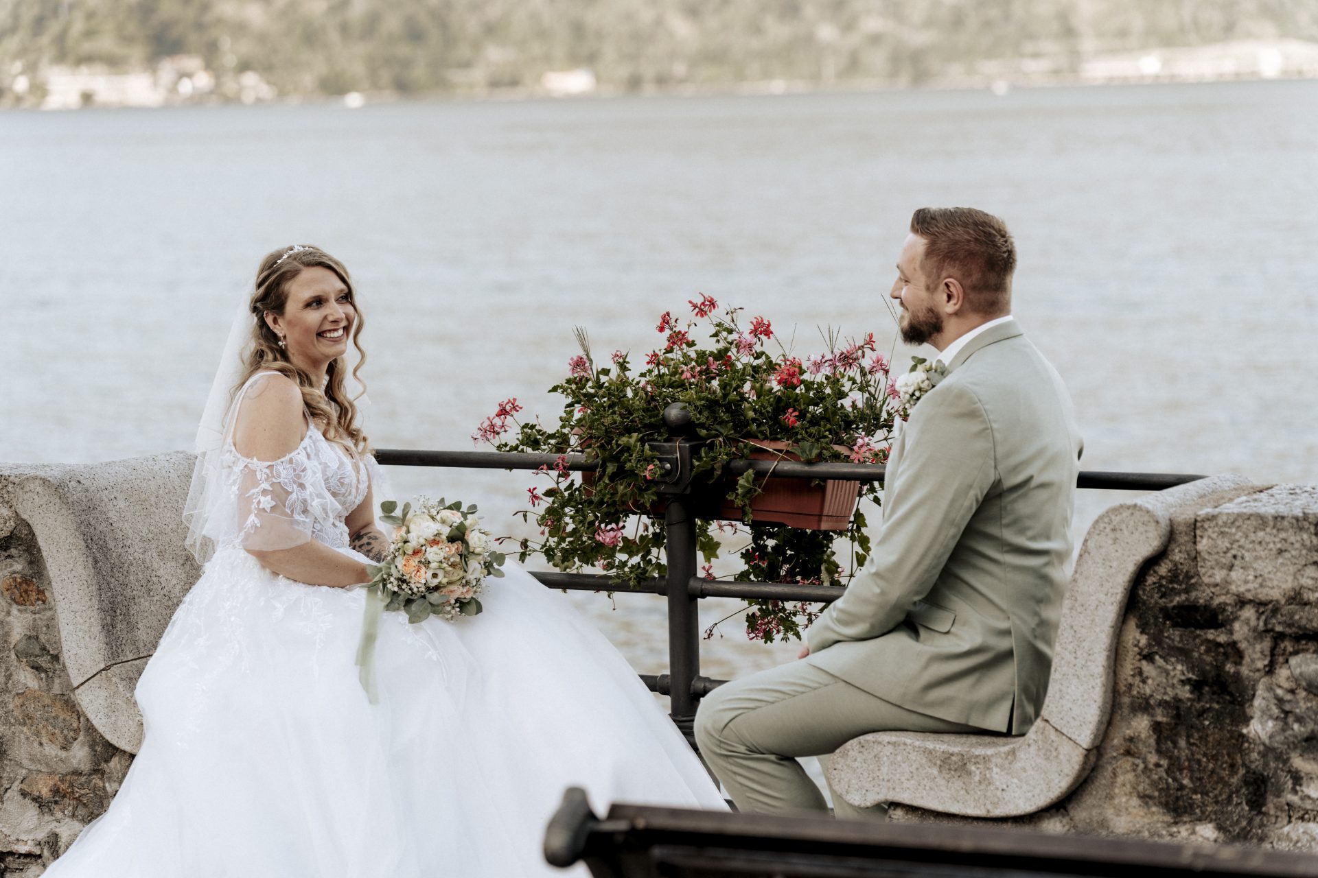 Braut und Bräutigam sitzen auf einer Steinbank mit Geranien am Lago Maggiore