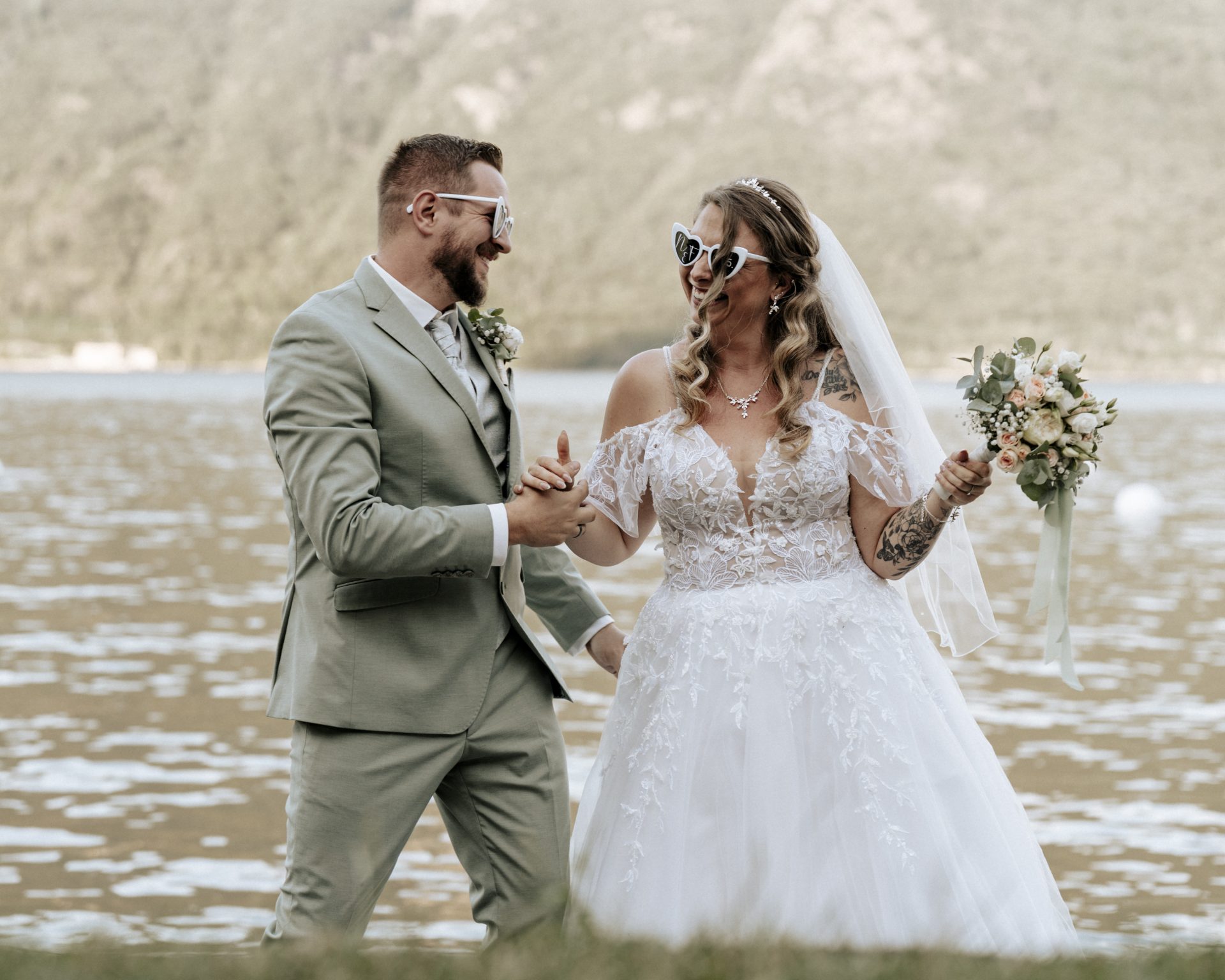 Bräutigam und Braut tanzen Hand in Hand mit Herzbrillen am Ufer