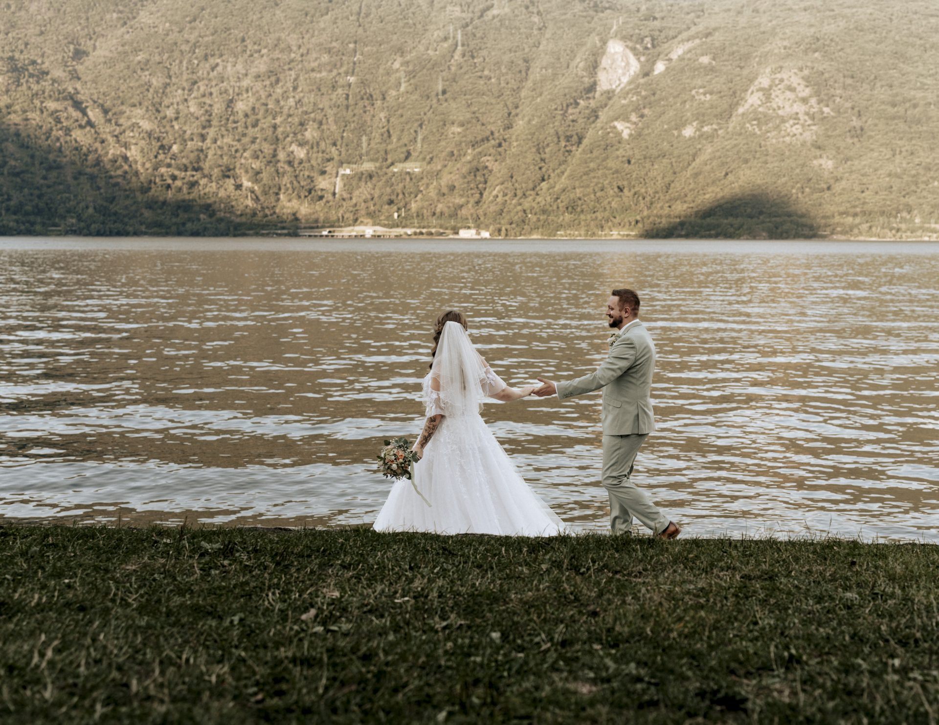 Brautpaar geht im Abendlicht Hand in Hand an der Uferwiese des Lago Maggiore