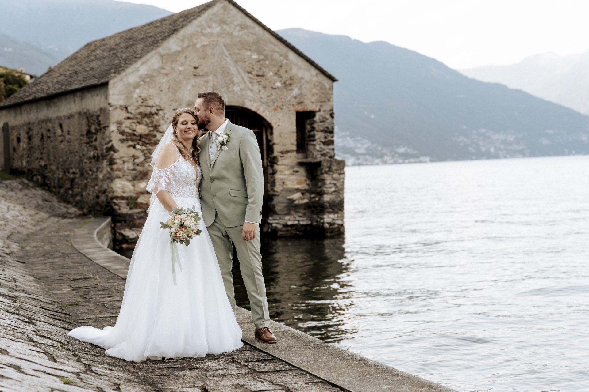 Brautpaar vor einem steinernen Bootshaus am Lago Maggiore mit Bergblick